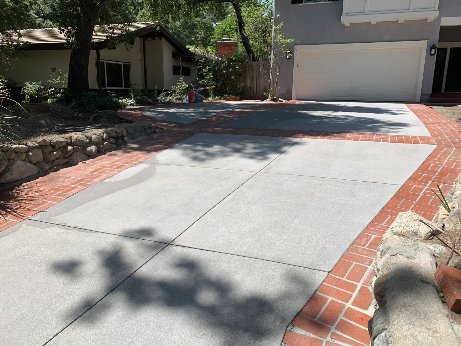 Concrete driveway with saw-cut joints and red herringbone brick border beside river rock retaining wall