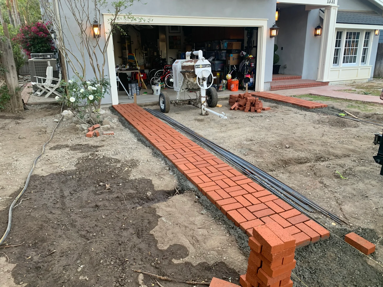 Herringbone red clay brick paver walkway under installation beside rebar and staged brick stacks on dirt