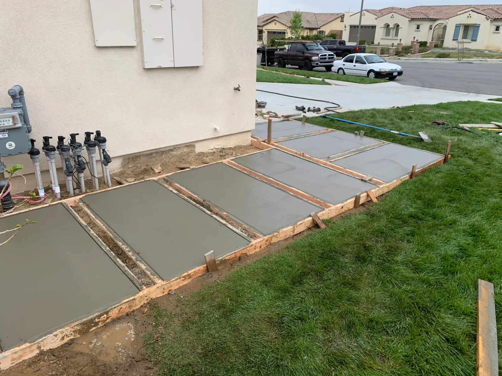 Freshly poured concrete slabs set within wood forms beside a stucco wall, with staked lumber and PVC irrigation pipes