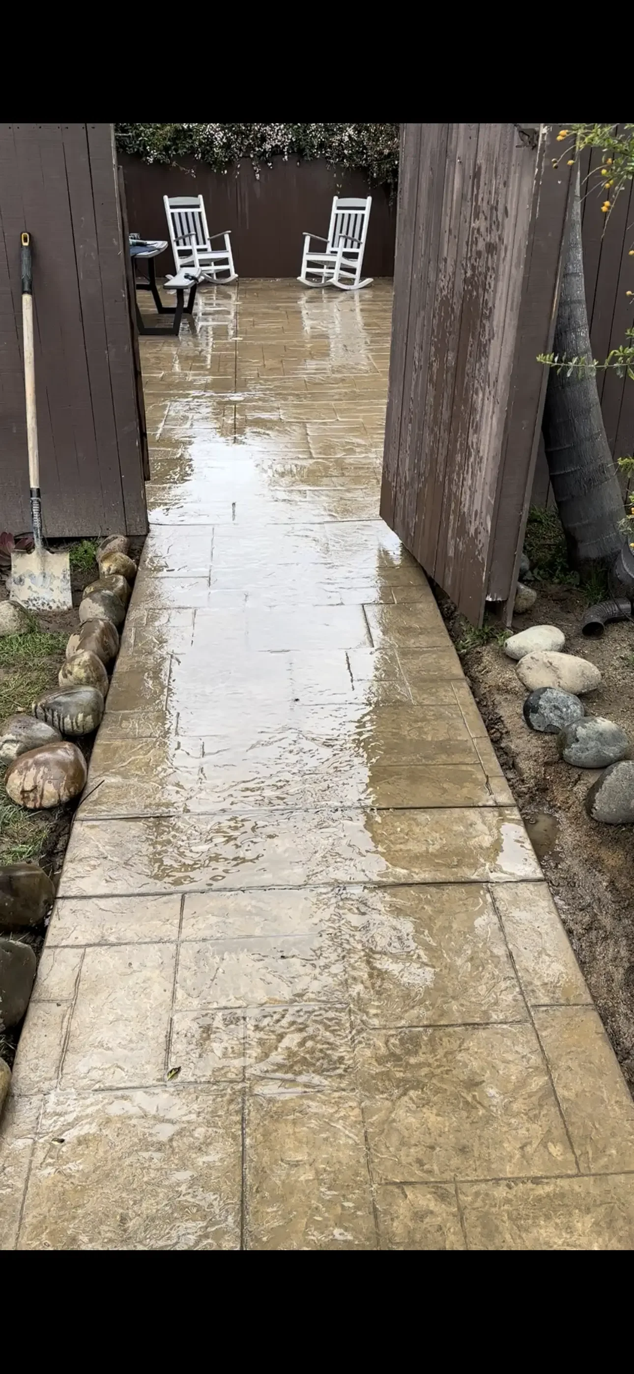 Stamped concrete walkway with slate-style texture and tan-brown color, bordered by river rock and leading to a patio beside wood fencing
