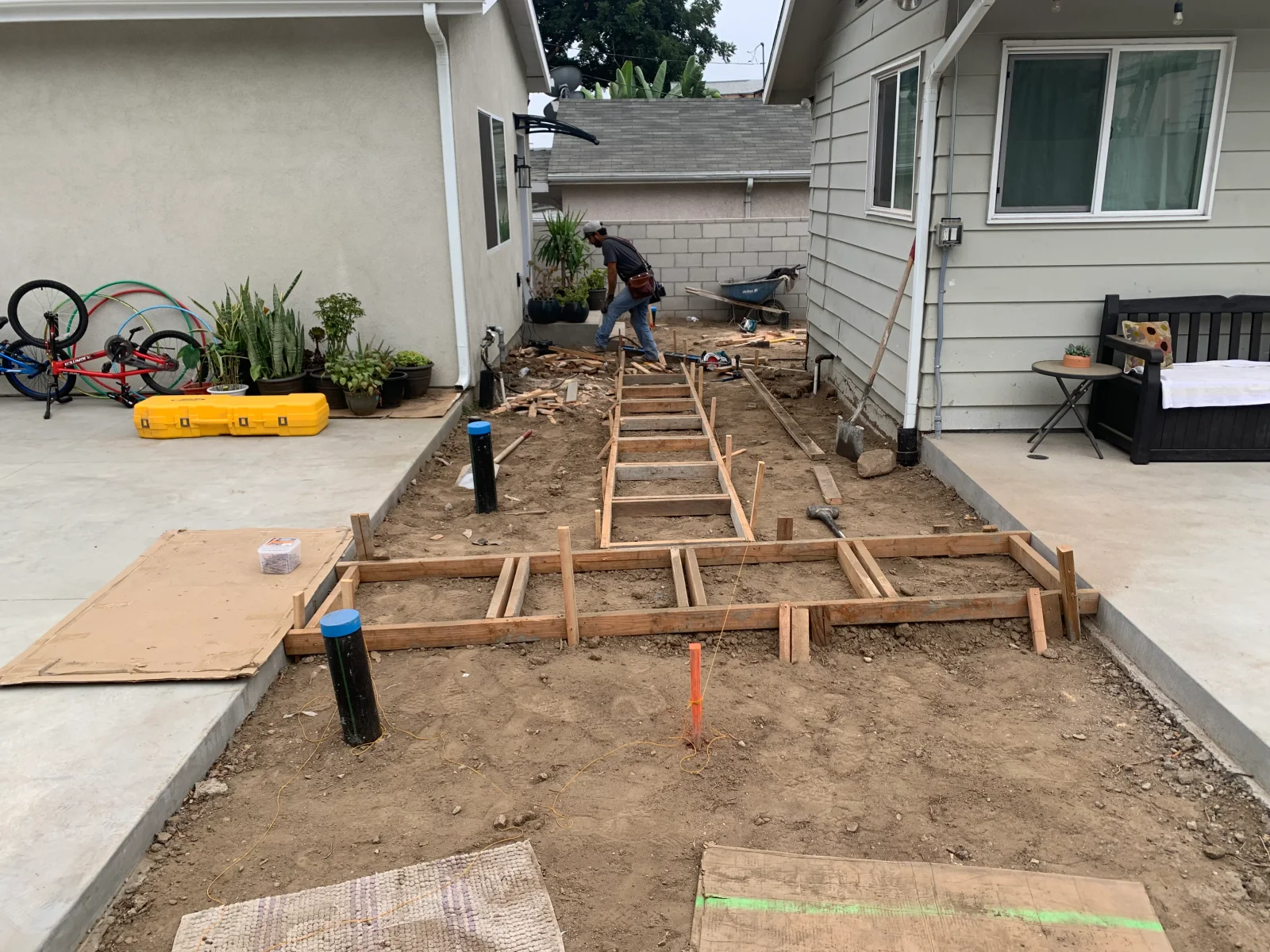Wood formwork and stakes set in dirt for a new concrete slab with utility conduits and string lines, beside existing concrete patio