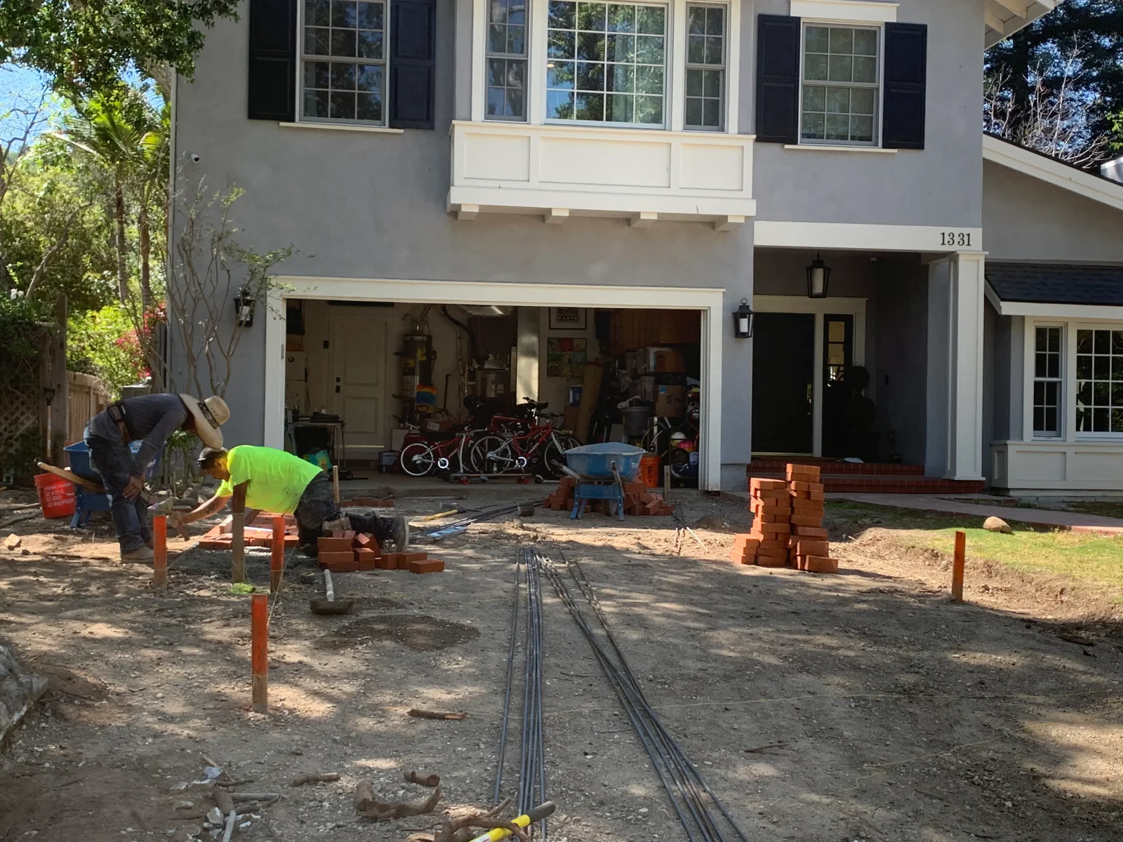 Workers laying red clay brick pavers on a graded driveway with stacked brick, rebar, and orange grade stakes