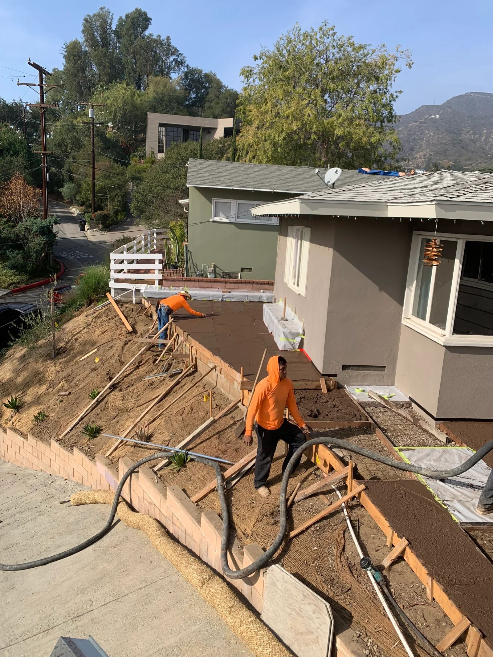 Crew placing and screeding fresh concrete slab within wood forms beside a stepped concrete block retaining wall on a slope
