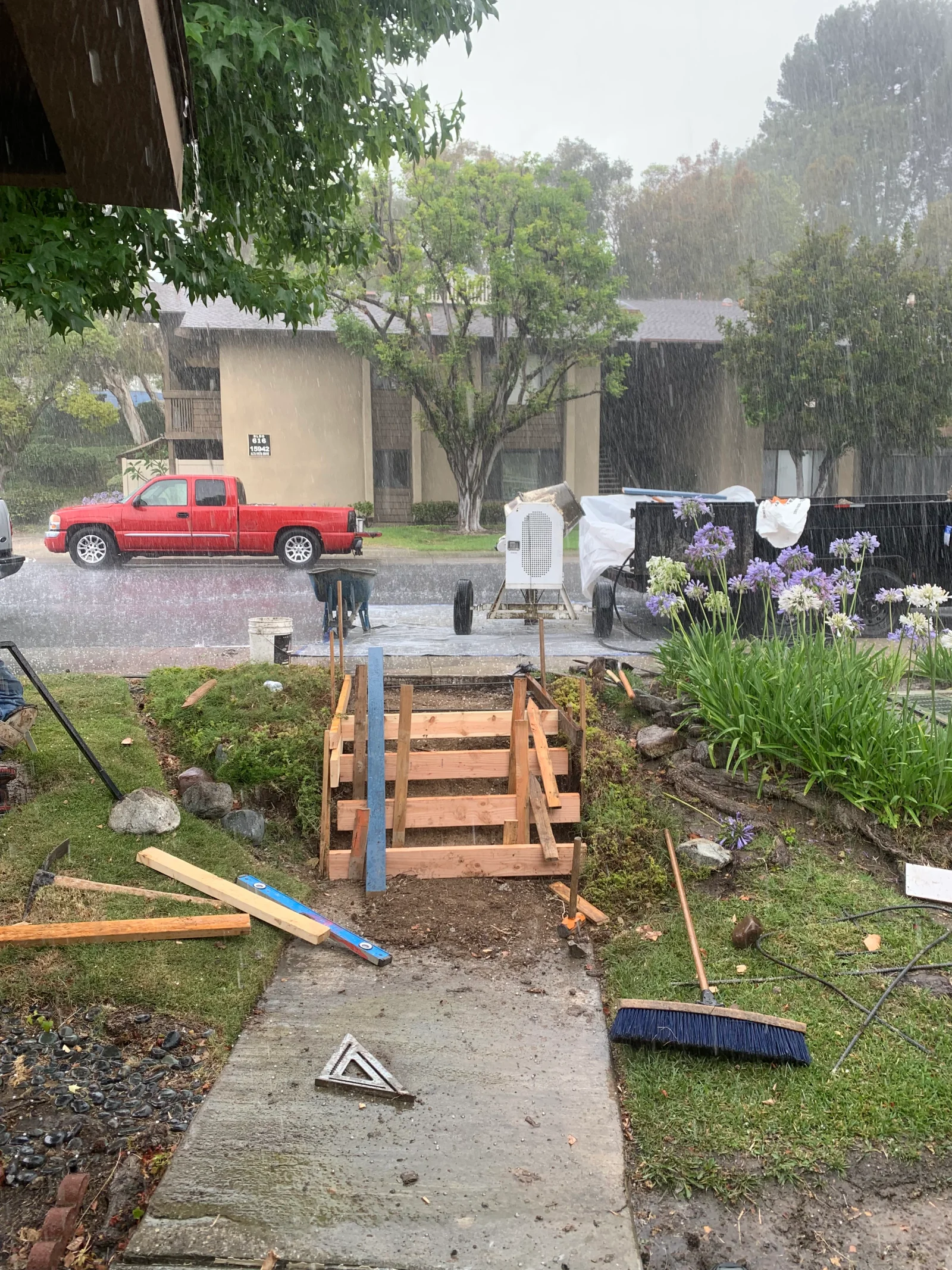 Wood forms and stakes set in a trench for a concrete walkway, with a concrete mixer and tools on site