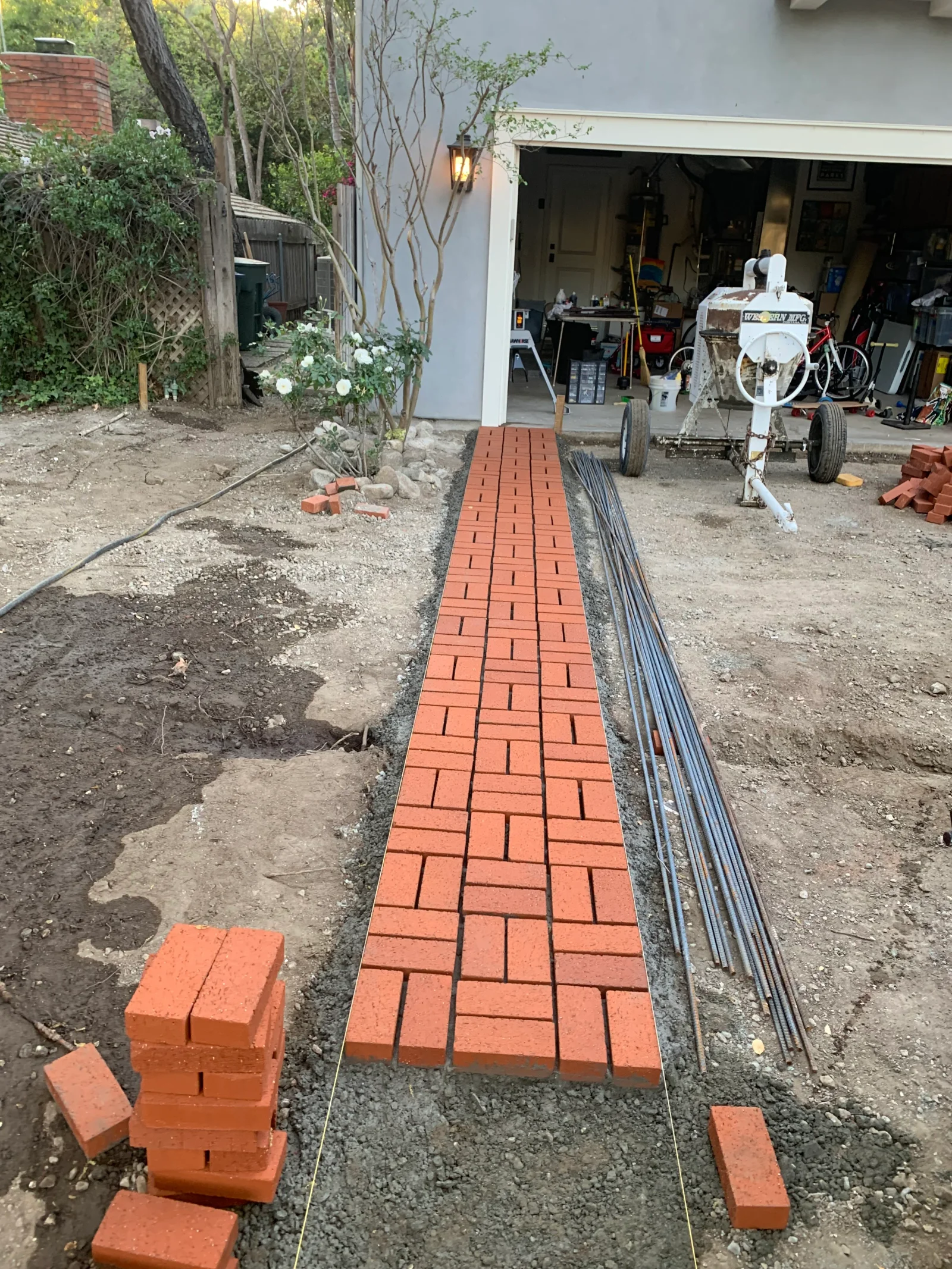 Herringbone red brick walkway being laid over gravel base beside stacked bricks and rebar near garage