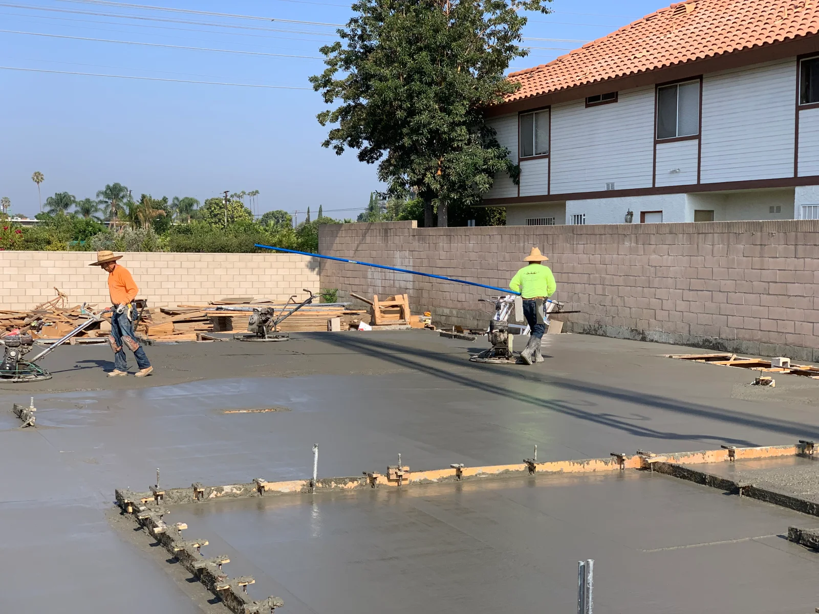 Crew finishing a fresh concrete slab with power trowel and long-handled float, wood forms and metal stakes at edges