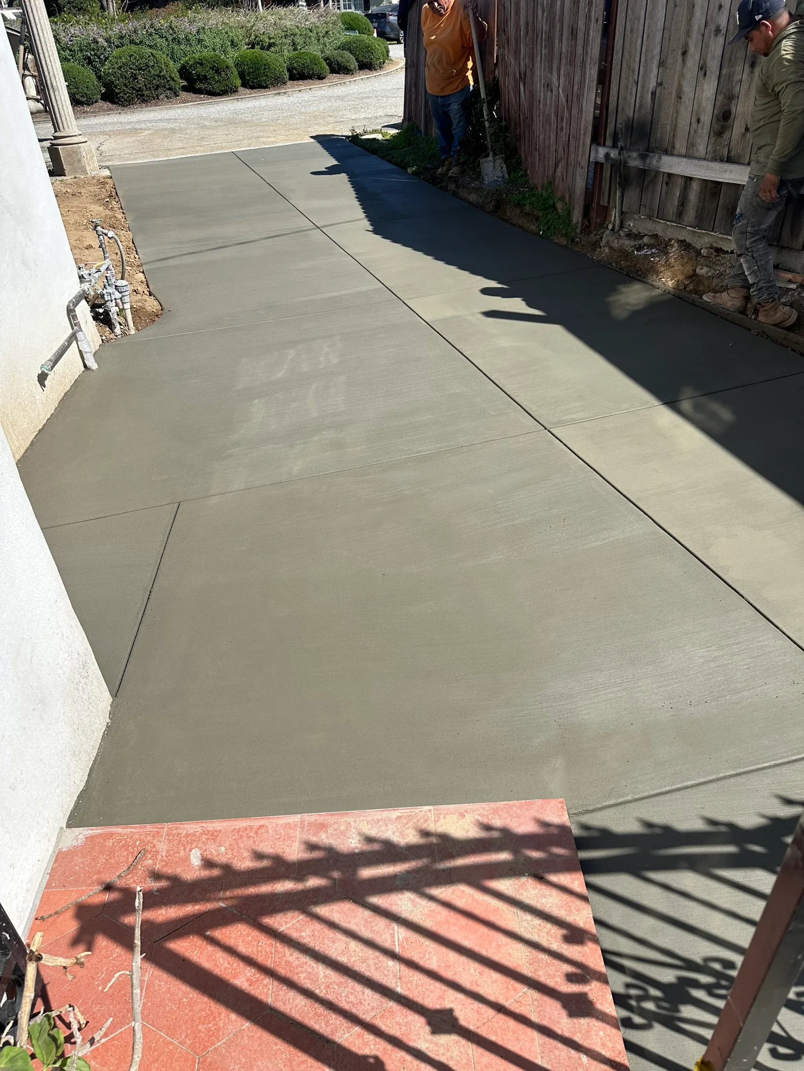 Freshly finished gray concrete driveway with saw-cut control joints beside a wood fence and stucco wall