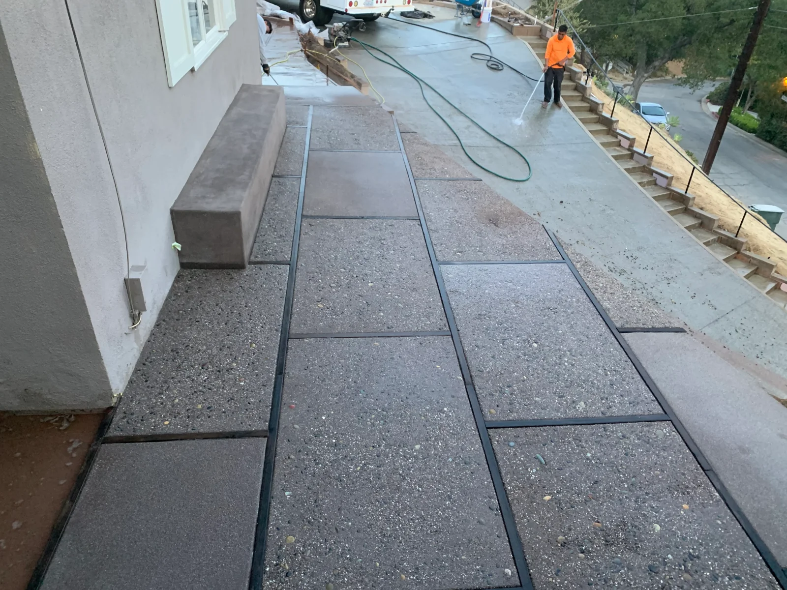 Exposed aggregate concrete walkway with dark divider strips beside a stucco wall and poured concrete bench