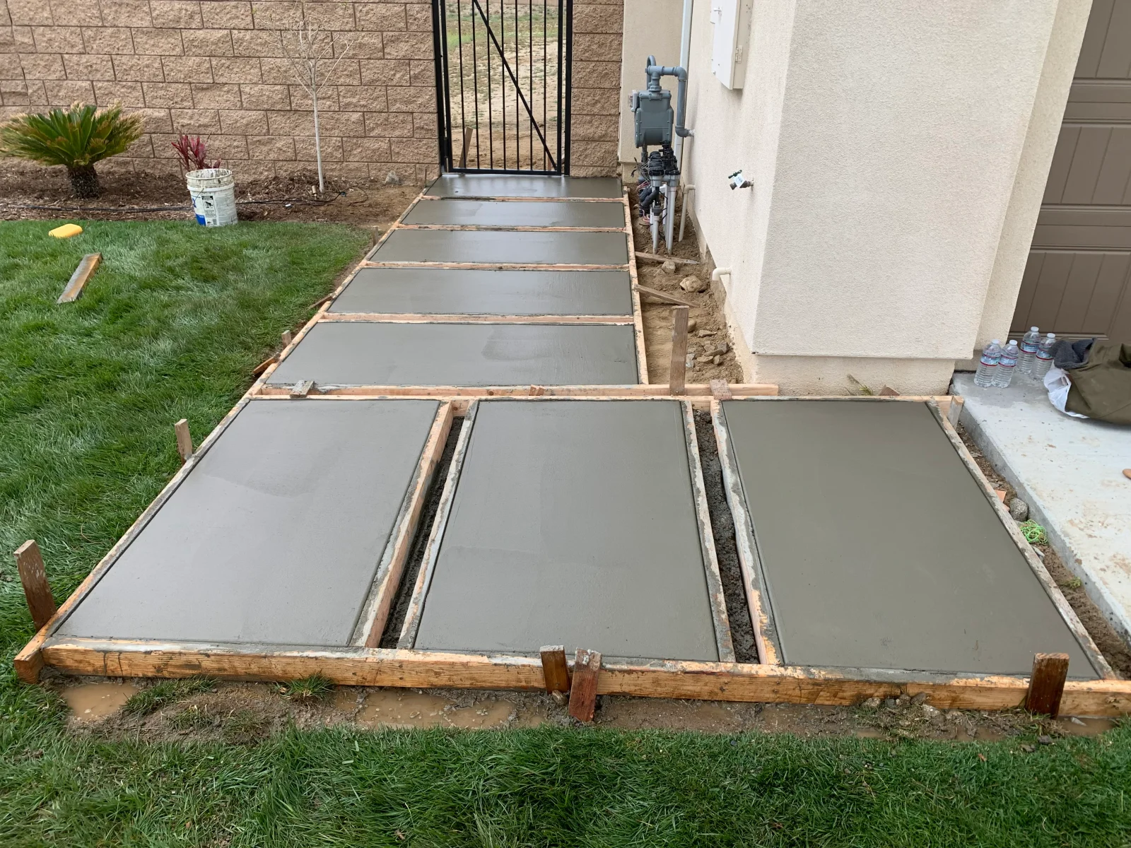 Freshly poured concrete walkway slabs held by wood forms beside stucco home wall and metal gate