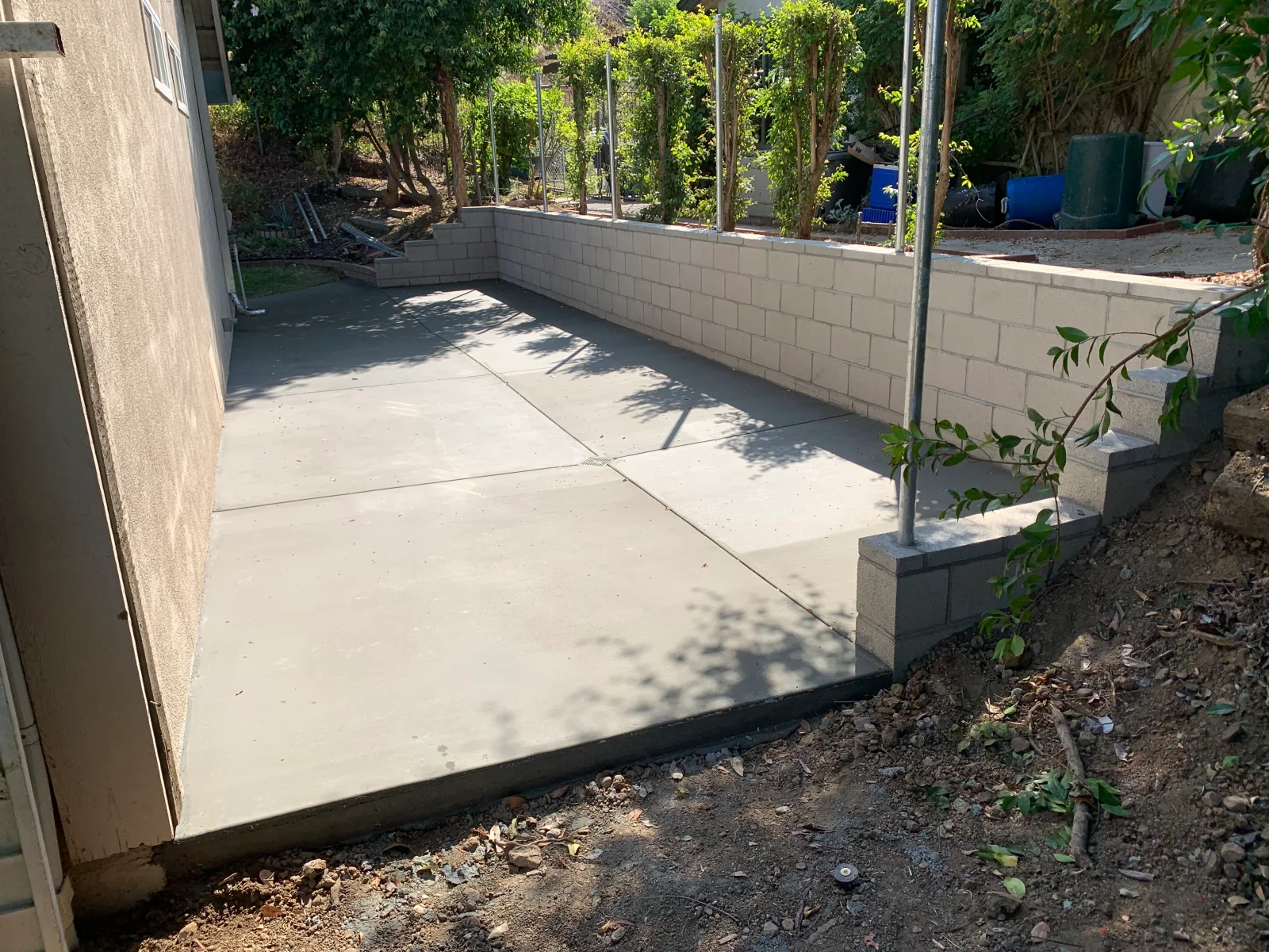 Newly poured smooth concrete patio with saw-cut control joints beside a concrete block retaining wall and steel posts