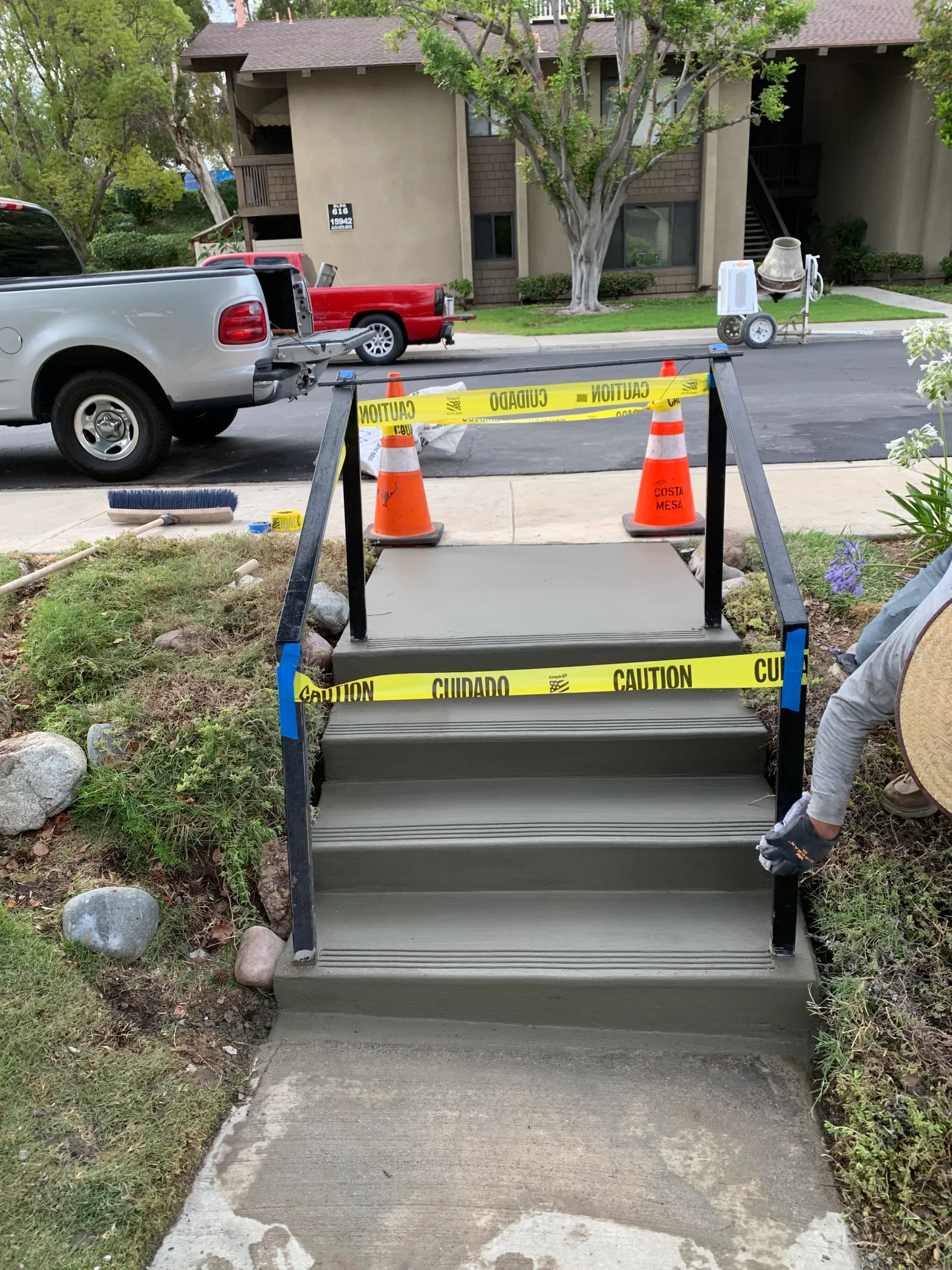 Newly poured gray concrete entry steps with grooved nosing and black steel handrails, marked off with caution tape and cones